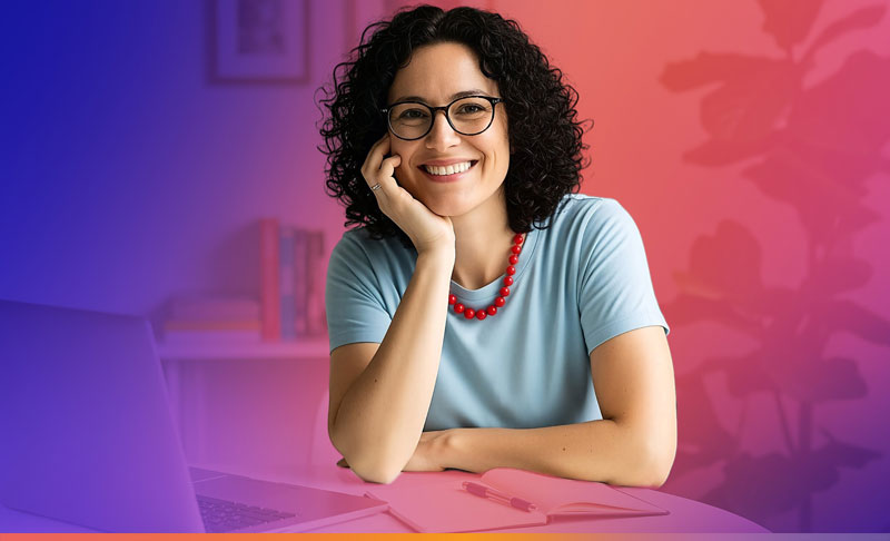 Frau mit Brille, die an einem Schreibtisch sitzt und lächelt, während sie auf einen Laptop schaut.