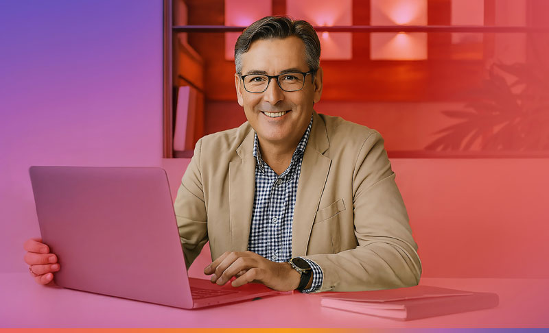 Mann in beigem Jackett, der an einem Laptop am Schreibtisch arbeitet und lächelt.