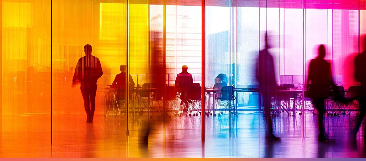 Zwei Personen arbeiten an Computern in einem Büro mit stark gesättigten Orange- und Rottönen, während andere vorbeigehen