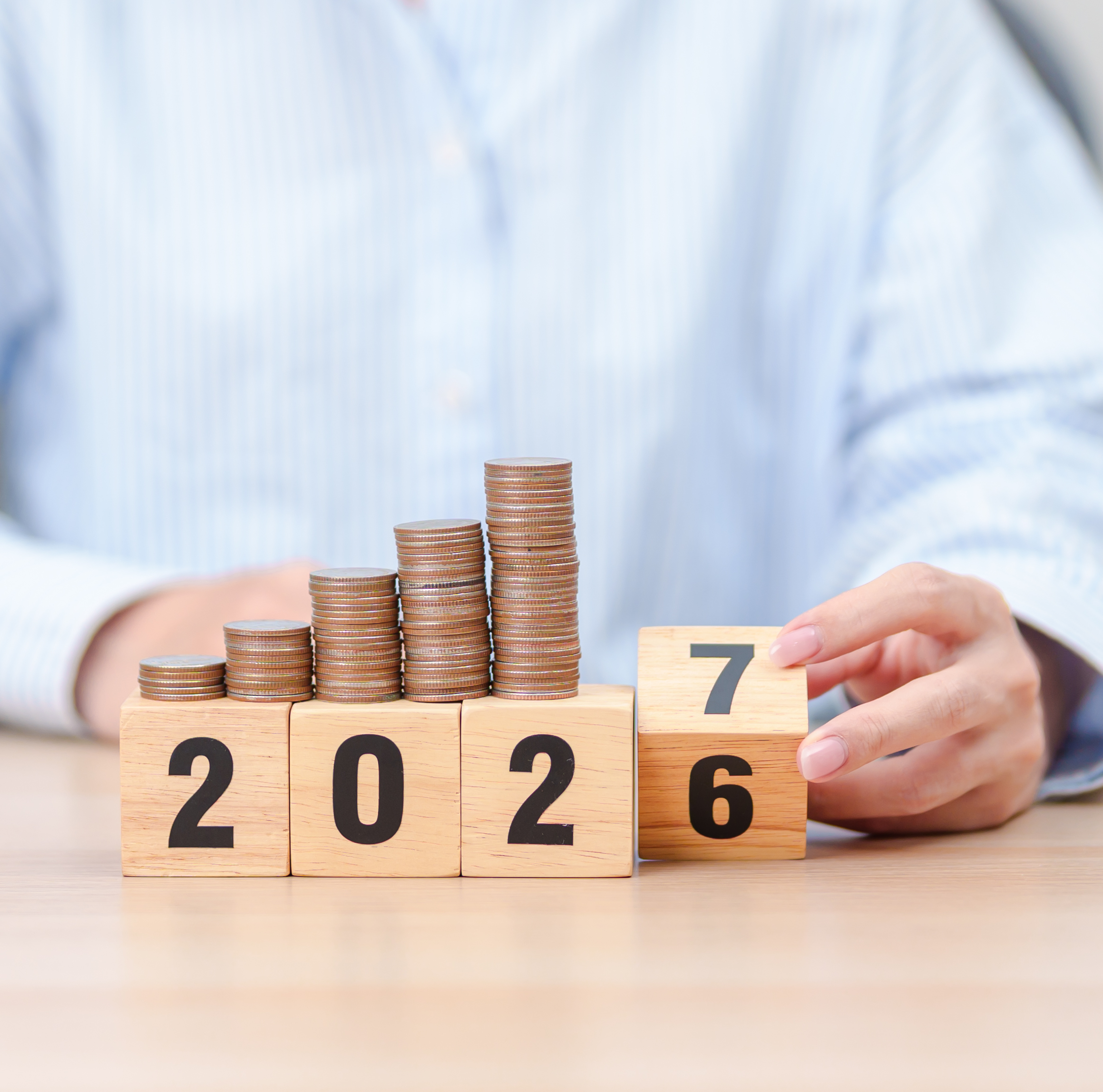 Symbolische Abbildung: Eine Frau sitzt vor 4 Zahlenwürfeln auf denen die Ziffern 2.0.2 zu lesen sind.  Sie dreht einen vierten Würfel von 6 auf 7 um, sodass aus 2026 nun 2027 wird. Auf den Würfeln sind Geldmünzen gestapelt.